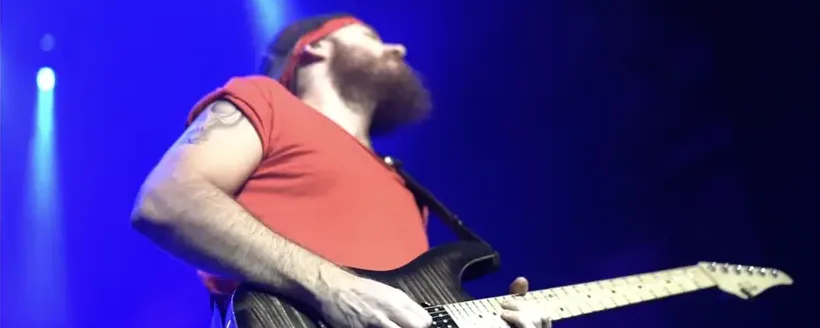 Ben Hartwell in a red T-shirt and headband playing electric guitar under blue stage lights