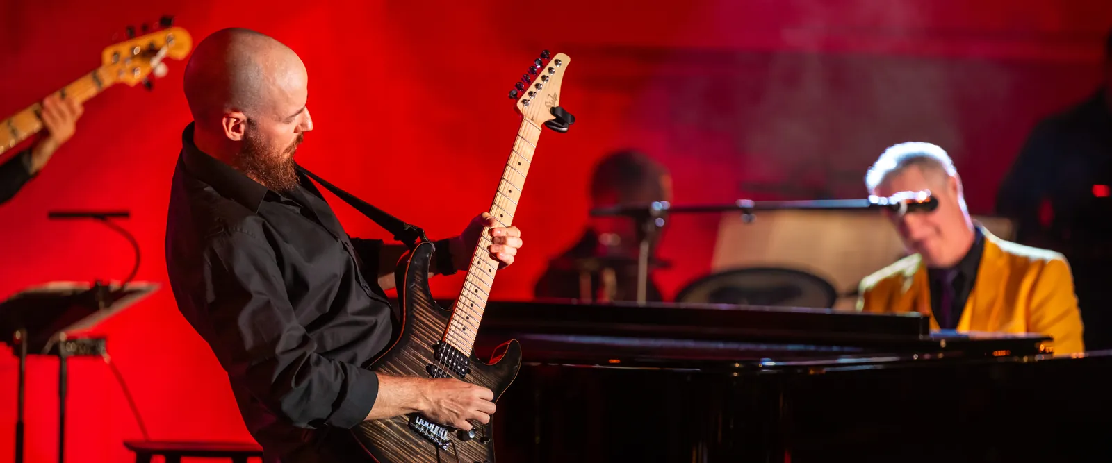 Ben Hartwell playing electric guitar on stage in front of a pianist at a grand piano, red backdrop