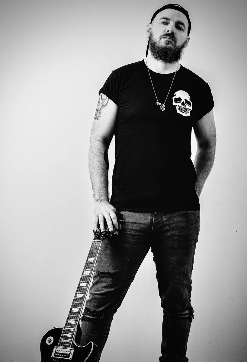 Black and white portrait of Ben Hartwell in a cap and skull T-shirt, resting his hand on a Les Paul guitar