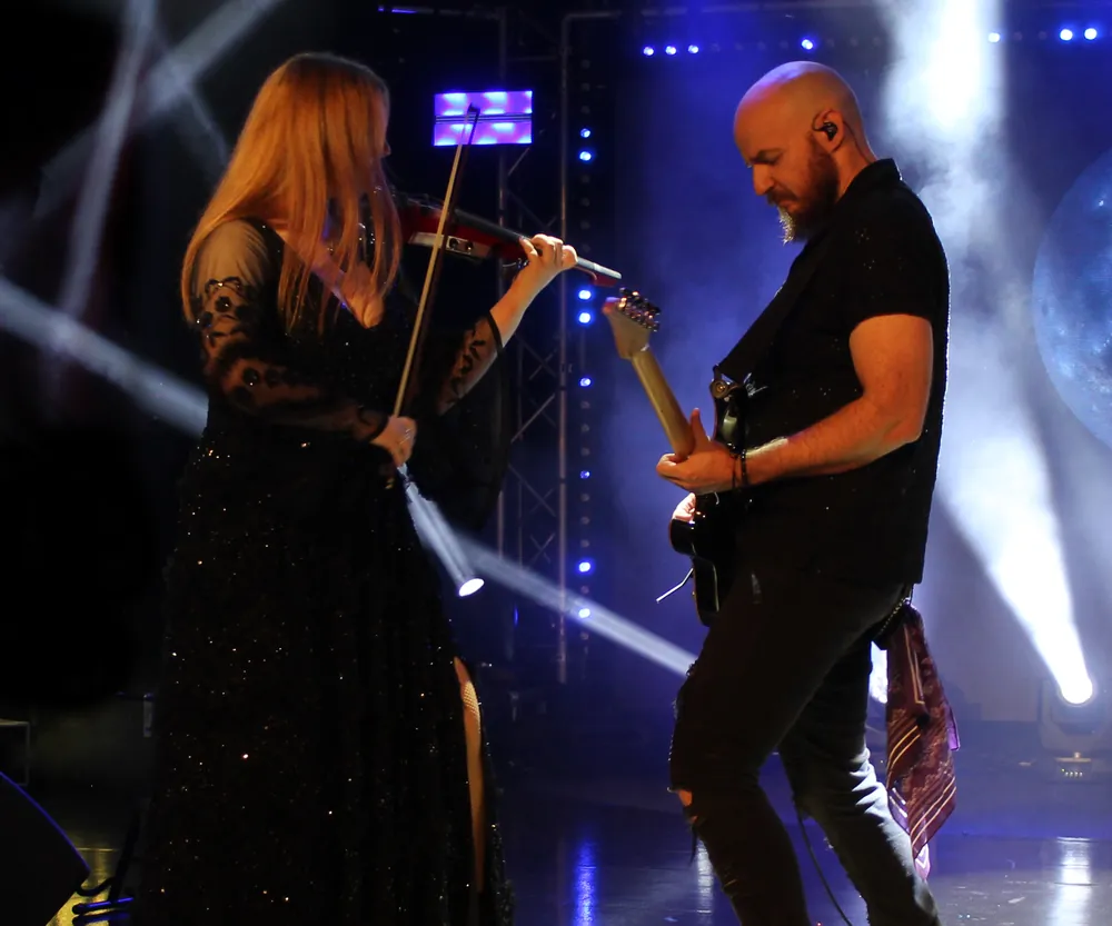 Ben Hartwell on electric guitar beside a violinist on a lit stage at a Stringstorm show